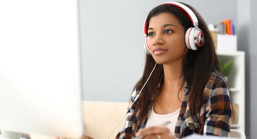 Woman of color sits at desk on computer wearing white headphones.