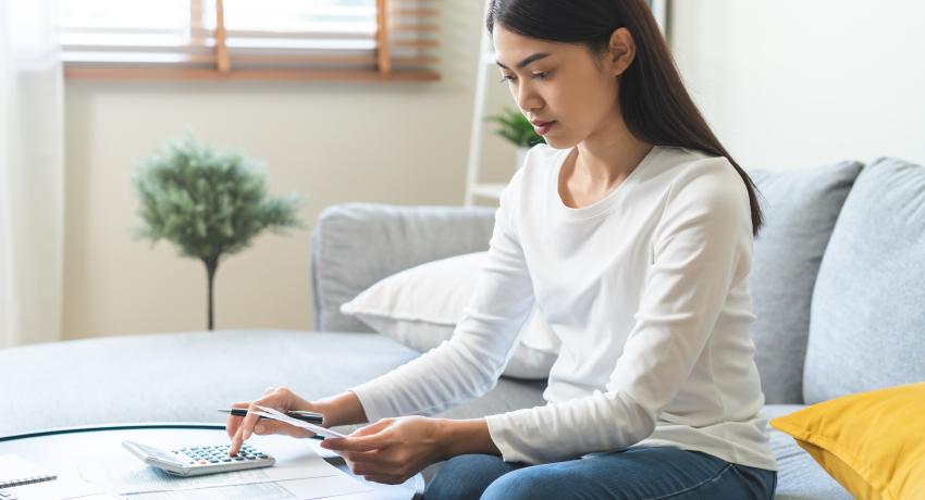 Young woman of color with long, black hair sitting on couch at desk typing on calculator.